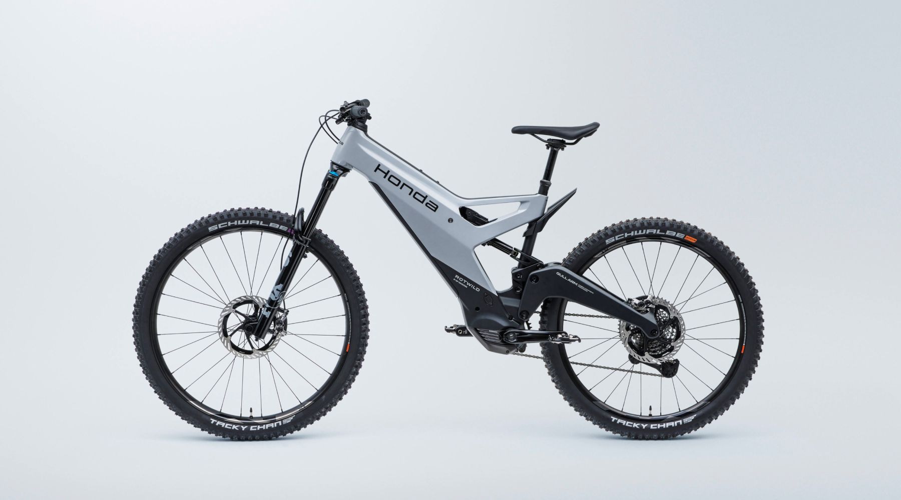 A side view of a Honda electric mountain bike with full suspension, featuring a light gray frame, black components, and large knobby tires. The bike has disc brakes and appears to be designed for off-road use.